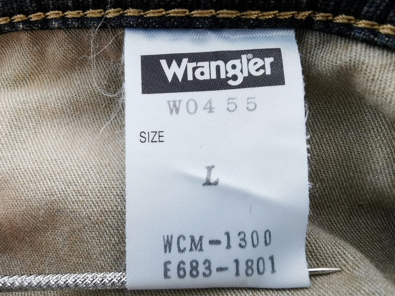 Peut inclure: Gros plan sur une &eacute;tiquette blanche avec le logo Wrangler et le texte "W04 55". L'&eacute;tiquette indique &eacute;galement la taille "L" et les codes "WCM-1300" et "E683-1801". L'&eacute;tiquette est fix&eacute;e &agrave; du tissu denim.