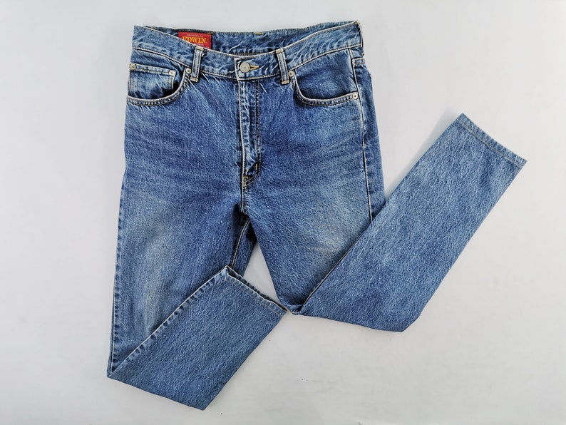 Puede incluir: Vaqueros de mezclilla azul claro con un dise&ntilde;o cl&aacute;sico de cinco bolsillos. Los vaqueros tienen cierre de bot&oacute;n y cremallera, trabillas para el cintur&oacute;n y un lavado descolorido. La marca "EDWIN" es visible en una etiqueta roja.