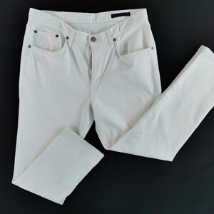May include: Cream-coloured denim jeans with a classic five-pocket design. The trousers feature a button and zip closure, belt loops, and a straight leg cut. The jeans are laid flat on a black surface.