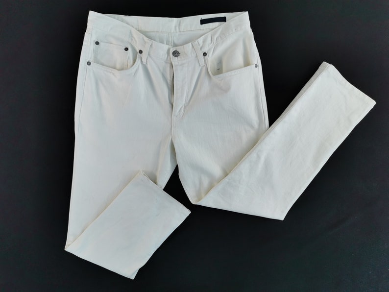 May include: Cream-coloured denim jeans with a classic five-pocket design. The trousers feature a button and zip closure, belt loops, and a straight leg cut. The jeans are laid flat on a black surface.