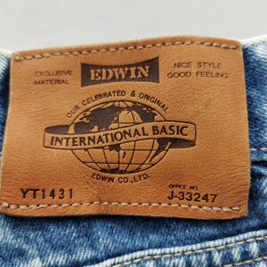 Puede incluir: Primer plano de una etiqueta de cuero marr&oacute;n cosida a unos vaqueros azules. La etiqueta dice "EDWIN INTERNATIONAL BASIC" con un gr&aacute;fico de globo. El texto adicional incluye "EXCLUSIVE MATERIAL" y "NICE STYLE GOOD FEELING". La etiqueta tambi&eacute;n tiene el texto "EDWIN CO. LTD." y los n&uacute;meros "YT1431" y "J-33247".