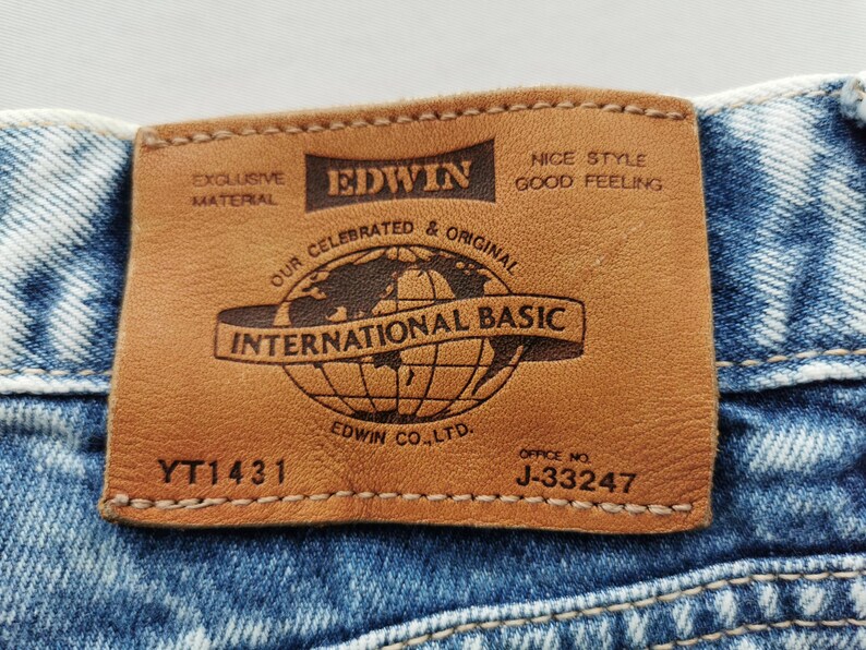 Puede incluir: Primer plano de una etiqueta de cuero marr&oacute;n cosida a unos vaqueros azules. La etiqueta dice "EDWIN INTERNATIONAL BASIC" con un gr&aacute;fico de globo. El texto adicional incluye "EXCLUSIVE MATERIAL" y "NICE STYLE GOOD FEELING". La etiqueta tambi&eacute;n tiene el texto "EDWIN CO. LTD." y los n&uacute;meros "YT1431" y "J-33247".