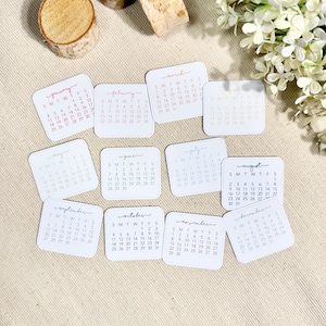 Puede incluir: Un conjunto de doce tarjetas cuadradas blancas, cada una con un mes del calendario de enero a diciembre impreso en un color distinto. Los meses est&aacute;n escritos en letra cursiva sobre una cuadr&iacute;cula de fechas, representando cada tarjeta un mes.