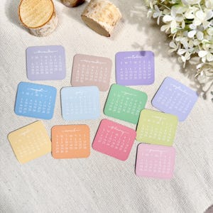 Puede incluir: Un conjunto de doce tarjetas de calendario cuadradas en varios colores pastel, cada una con un mes espec&iacute;fico de enero a diciembre con texto blanco impreso para los d&iacute;as y fechas, dispuestas en una cuadr&iacute;cula sobre una tela texturizada color crema.