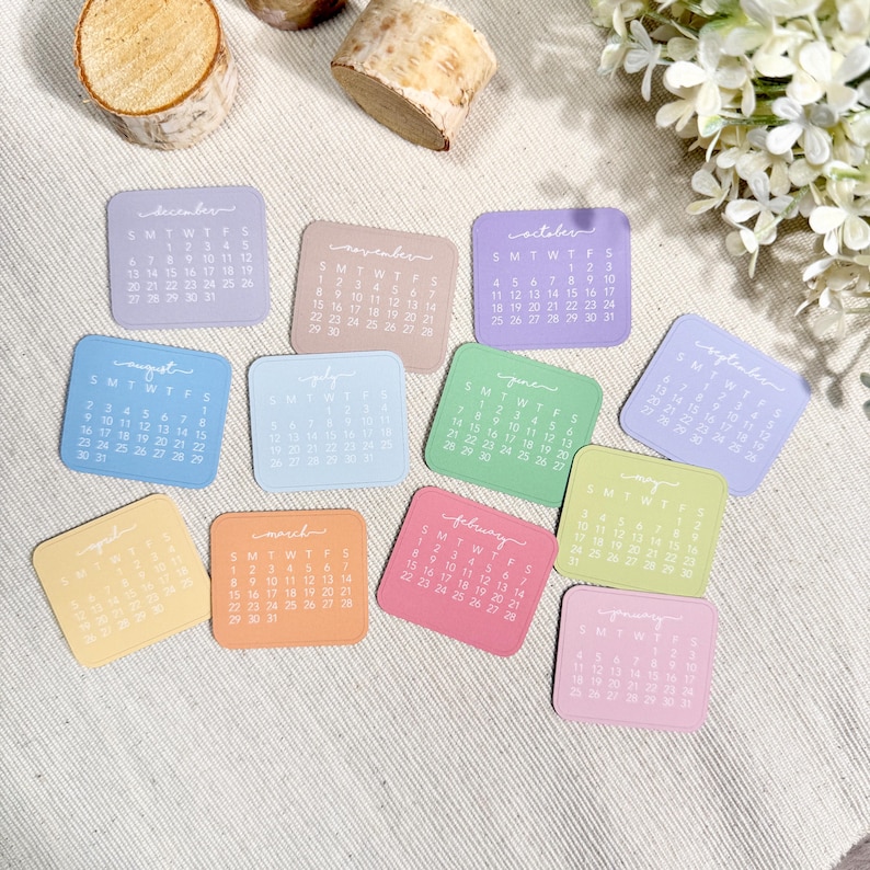 Puede incluir: Un conjunto de doce tarjetas de calendario cuadradas en varios colores pastel, cada una con un mes espec&iacute;fico de enero a diciembre con texto blanco impreso para los d&iacute;as y fechas, dispuestas en una cuadr&iacute;cula sobre una tela texturizada color crema.