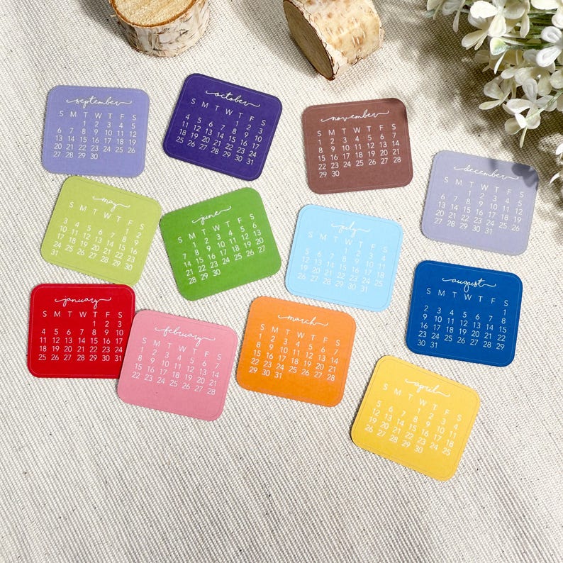 Puede incluir: Un conjunto de doce tarjetas de calendario cuadradas, cada una con un mes diferente del a&ntilde;o en un color &uacute;nico. Las tarjetas muestran el nombre del mes en letra cursiva en la parte superior, seguido de una cuadr&iacute;cula de d&iacute;as y fechas.