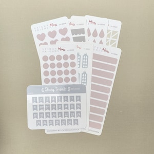 Può includere: Un set di 10 fogli di adesivi con vari disegni, tra cui cuori, cerchi, gocce e bandiere. Gli adesivi sono in tonalità di rosa e grigio. I fogli sono etichettati "Sticky Friends Mini" e hanno un codice prodotto.
