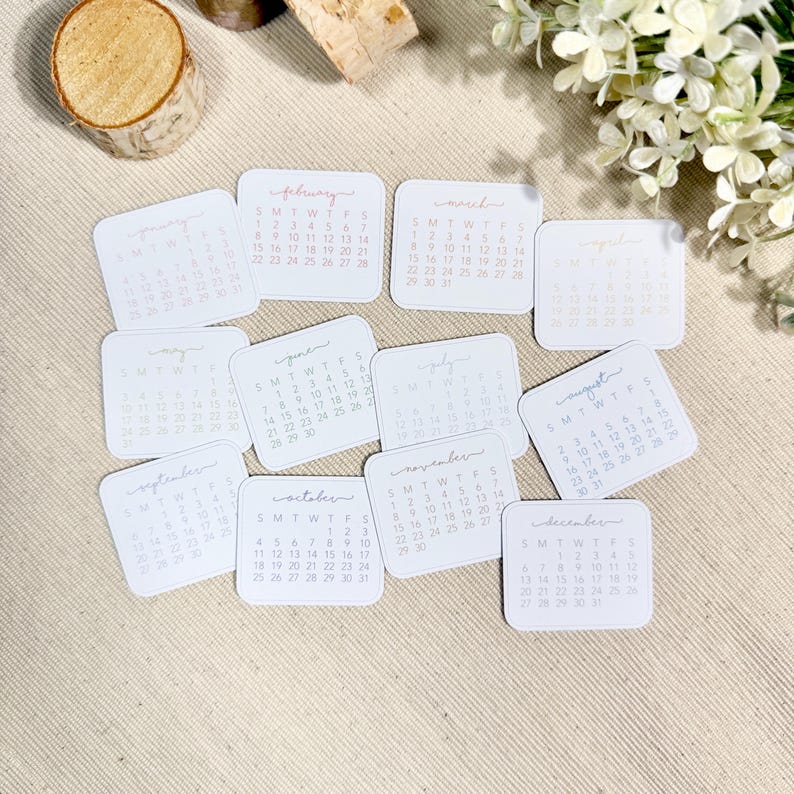 Puede incluir: Un conjunto de doce tarjetas cuadradas de papel blanco, cada una con un mes de calendario impreso de enero a diciembre con texto negro y un encabezado de mes en cursiva. Las tarjetas est&aacute;n dispuestas en un grupo sobre una superficie beige texturizada.
