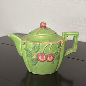 Könnte beinhalten: Eine hellgrüne Keramik-Teekanne mit einem erhabenen Reliefdesign aus roten Kirschen und grünen Blättern auf dem Körper. Die Teekanne hat einen passenden Deckel mit einem kleinen roten Knauf, einen geometrischen Griff und einen spitzen Ausguss.