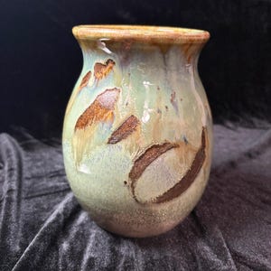 May include: A ceramic vase with a rounded body and a flared rim, featuring a variegated glaze in shades of sage green, cream, and brown. The surface displays textured, dark brown patches and a smooth, glossy finish throughout the piece.
