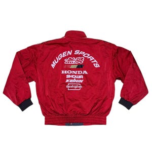May include: A red Mugen Sports jacket with white logos and text. The jacket has a ribbed waist and cuffs, and the back displays Honda, Showa, Keihin, and denshigiken logos. A vintage racing-inspired design.
