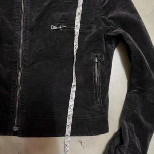 May include: A dark grey corduroy jacket with a zip closure and two zipped pockets. The jacket has a tailored fit and is being measured with a tape measure. The jacket's length is approximately 51 cm.