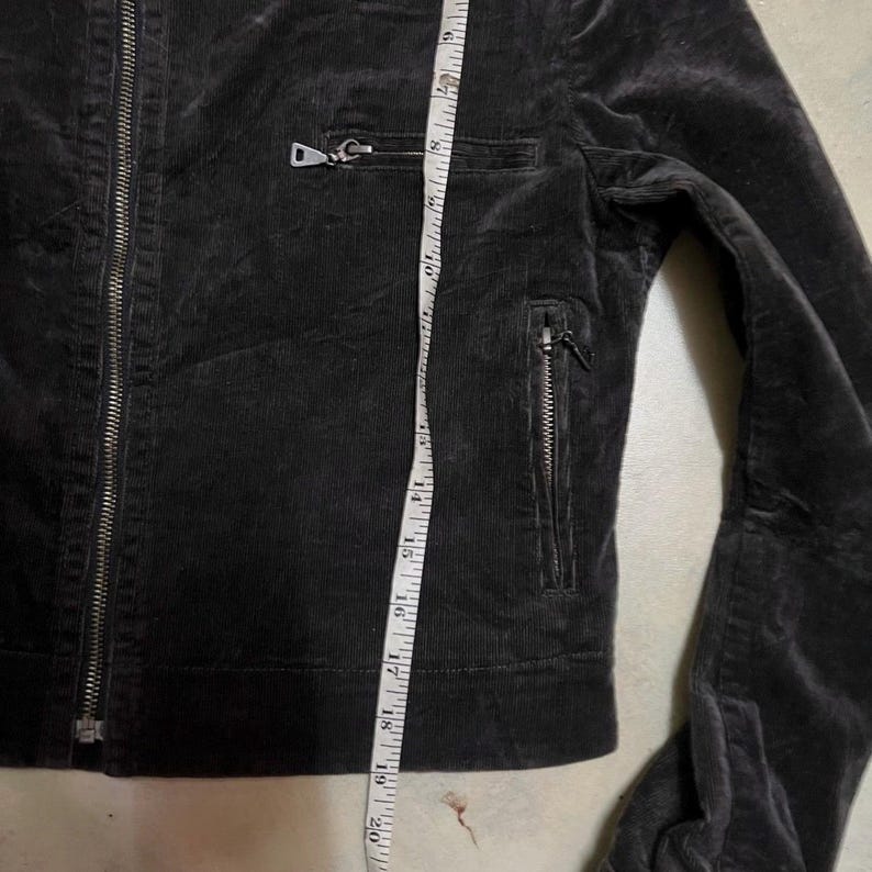 May include: A dark grey corduroy jacket with a zip closure and two zipped pockets. The jacket has a tailored fit and is being measured with a tape measure. The jacket's length is approximately 51 cm.