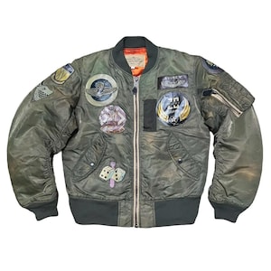 Op de afbeelding: Een groene bomberjack met meerdere patches, waaronder een dobbelsteenpatch, een patch met de tekst "20th AIR FORCE" en een patch met de tekst "AVIREX".
