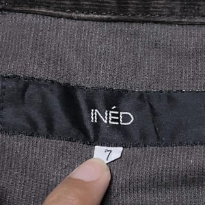 May include: Close-up of dark brown corduroy fabric with a black label. The label reads "IN&Eacute;D" in white capital letters. Below the label is a small white tag with the number "7" printed on it.