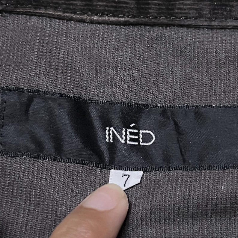 May include: Close-up of dark brown corduroy fabric with a black label. The label reads "IN&Eacute;D" in white capital letters. Below the label is a small white tag with the number "7" printed on it.