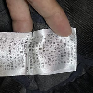 May include: A close-up of a white label with Japanese text and the numbers 331/331OKH. The label is attached to a dark grey fabric. The text on the label provides care instructions and warnings about colour transfer and changes in lustre.