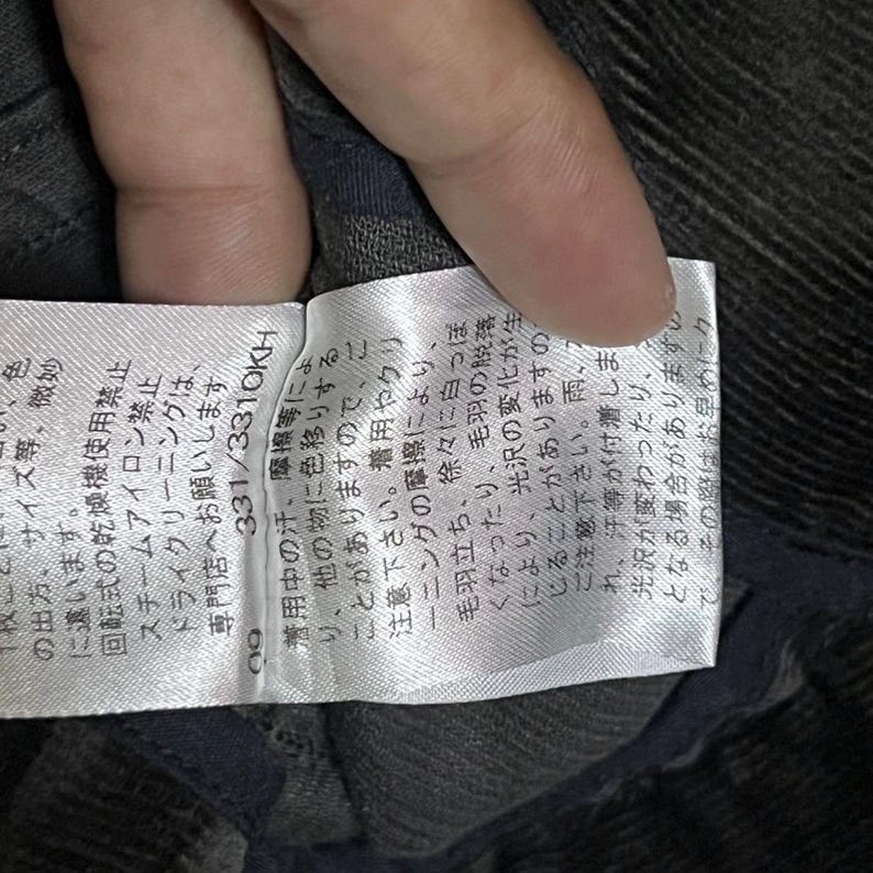 May include: A close-up of a white label with Japanese text and the numbers 331/331OKH. The label is attached to a dark grey fabric. The text on the label provides care instructions and warnings about colour transfer and changes in lustre.