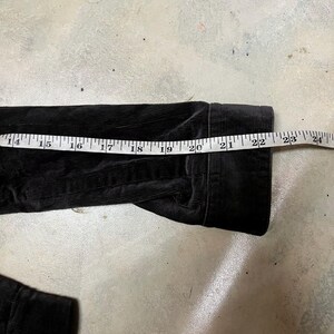 May include: Black corduroy jacket sleeve with a measuring tape showing a length of approximately 56 cm. The jacket has a ribbed texture and is lying on a concrete surface.