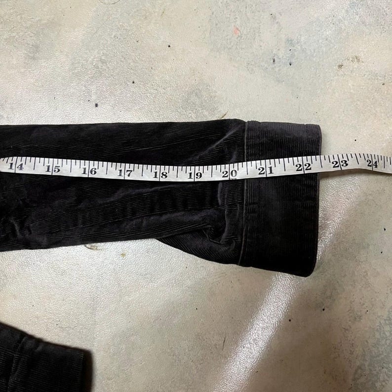 May include: Black corduroy jacket sleeve with a measuring tape showing a length of approximately 56 cm. The jacket has a ribbed texture and is lying on a concrete surface.