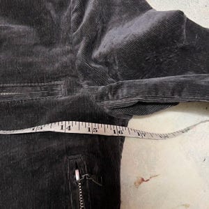 May include: Close-up of a dark grey corduroy jacket with a zip pocket and a measuring tape showing a length of approximately 35.5 to 40.6 cm. The jacket has a ribbed texture.