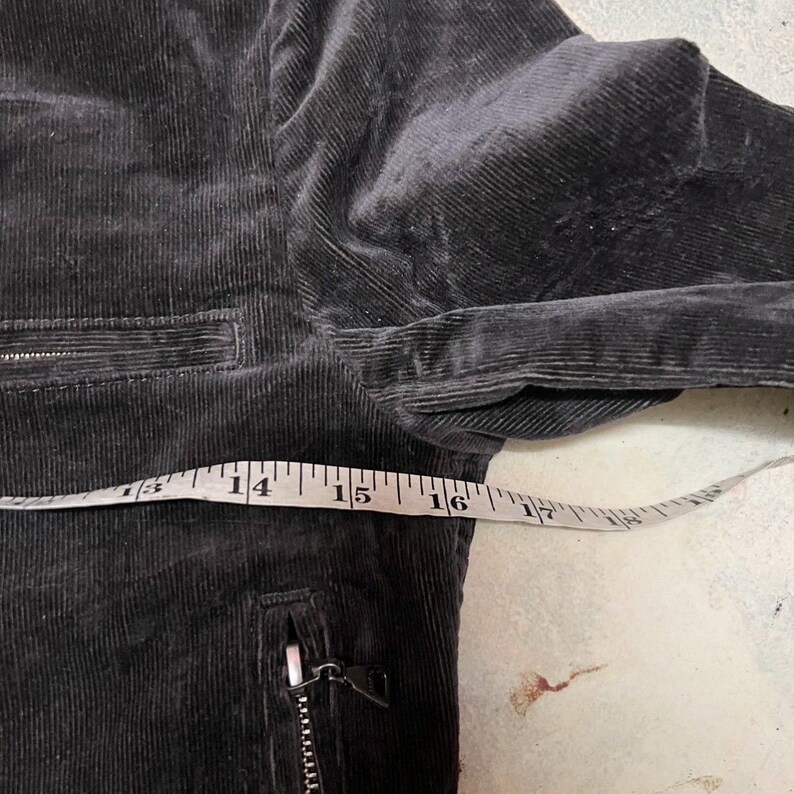 May include: Close-up of a dark grey corduroy jacket with a zip pocket and a measuring tape showing a length of approximately 35.5 to 40.6 cm. The jacket has a ribbed texture.