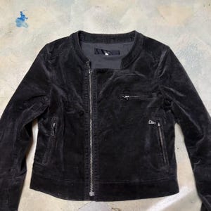May include: A black velvet jacket featuring a round collar and an asymmetrical zip. It has two zipped pockets at the front and one on the chest. The long-sleeved jacket is a deep black colour, suitable for various occasions.