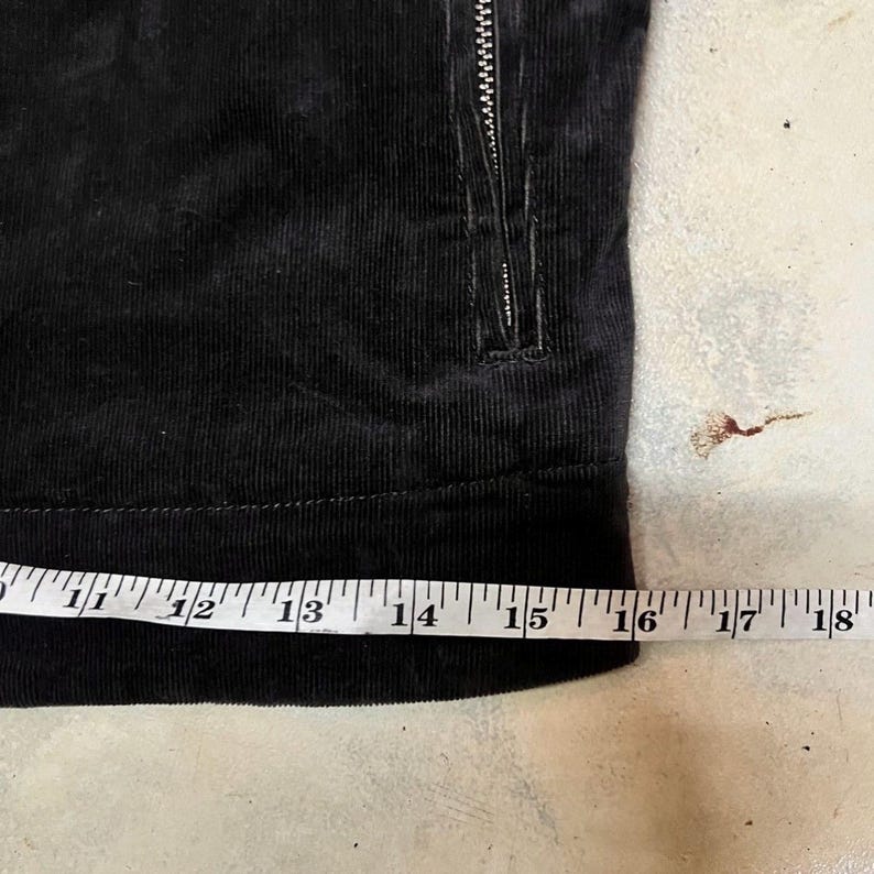 May include: Close-up of black corduroy fabric with a zipper. A measuring tape shows the fabric is approximately 40 cm wide. The corduroy has a ribbed texture.