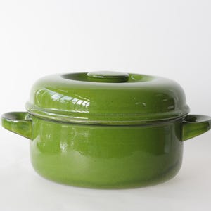 Può includere: Una pentola da cucina in ceramica verde con coperchio e due manici laterali. La pentola ha una finitura lucida e una forma arrotondata. Il coperchio ha un piccolo pomello sulla parte superiore. La pentola è progettata per cucinare.
