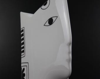 Könnte beinhalten: Eine weiße Keramikvase mit einem stilisierten Gesichtsdesign. Die Vase zeigt ein Profil mit Nase und Augendetails sowie schwarzen geometrischen Mustern. Die Vase hat eine moderne, künstlerische Ästhetik, geeignet für die Inneneinrichtung.