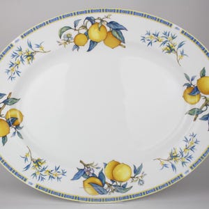Könnte beinhalten: Ovale Servierplatte mit weißem Hintergrund, verziert mit einem Zitronen- und Blumendesign. Der Rand weist ein blau-gelbes geometrisches Muster auf. Das Design der Platte umfasst gelbe Zitronen, blaue Blätter und zarte gelbe Blüten.