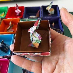 Può includere: Un piccolo coniglio di origami colorato in una scatola con motivo floreale. Il coniglio è fatto di carta e ha un design floreale. La scatola è marrone con un motivo floreale.