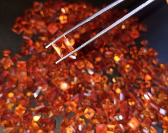 May include: A collection of small, square-cut, orange-colored gemstones. One gemstone is held by tweezers, showcasing its vibrant color and clarity. The gemstones are scattered on a dark surface, creating a sparkling effect.