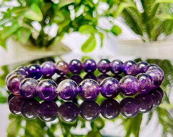 May include: A bracelet featuring polished, round amethyst beads. The purple beads are strung together, reflecting light. The bracelet is displayed on a reflective surface, with green foliage in the background. The beads are approximately 1 cm in diameter.