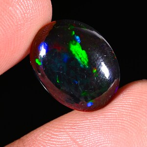 Könnte beinhalten: Ein ovaler schwarzer Opal-Edelstein mit einer polierten, glatten Oberfläche, der lebhafte Blitze in Neongrün, Elektroblau und subtilem Rot im gesamten dunklen, durchscheinenden Körper des Steins vor einem schwarzen Hintergrund zeigt.