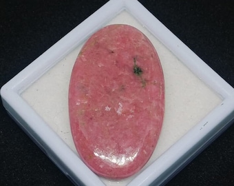 May include: A pink oval-shaped gemstone with a smooth, polished surface. The gemstone has a light pink color with darker pink and white inclusions. The gemstone is displayed in a white square box.