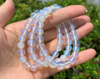 May include: A collection of opalescent beaded bracelets. The beads are round and translucent, with a soft, milky white color and subtle blue and purple hues. The bracelets are held in a hand against a blurred green background.