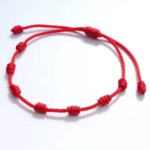 May include: A bright red cord bracelet featuring a series of evenly spaced, hand-tied knots along the length of the string. The bracelet is constructed from a twisted rope material and includes two loose ends that are also finished with small knots.