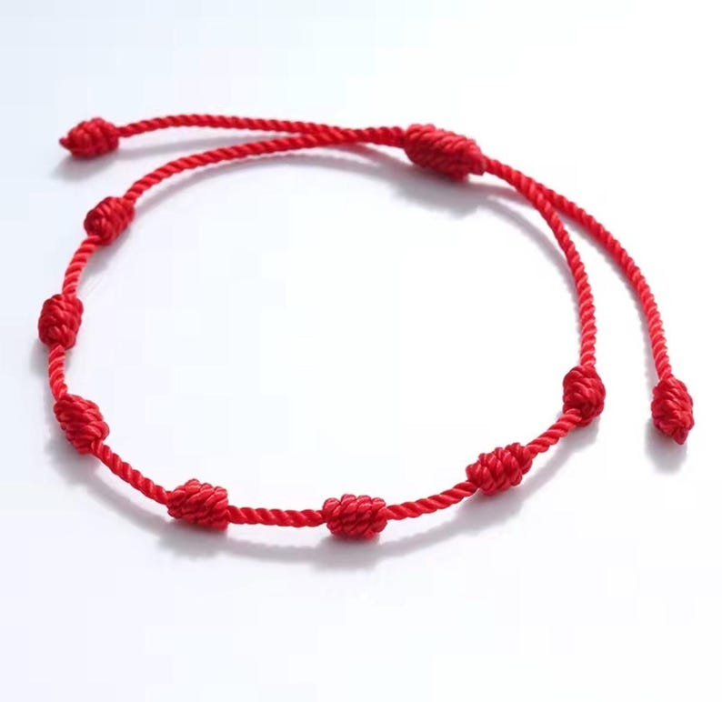 May include: A bright red cord bracelet featuring a series of evenly spaced, hand-tied knots along the length of the string. The bracelet is constructed from a twisted rope material and includes two loose ends that are also finished with small knots.