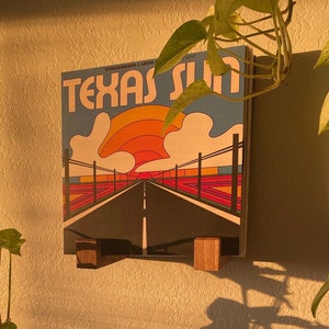 May include: A poster titled "Texas Sun" with a road leading to a sunset. The poster is displayed on a wall with wooden supports. A black amplifier with silver knobs and a small green speaker are in the foreground. A potted plant is on top of the amplifier.