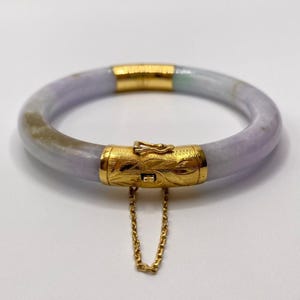 Puede incluir: Un brazalete de jade lavanda con cierre y cadena de oro. El brazalete está hecho de jade natural y tiene un diseño delicado y femenino.