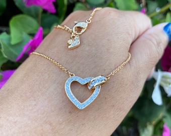 Pode incluir: Um colar dourado com um pingente em forma de coração. O pingente é adornado com cristais transparentes e tem um pequeno charme com cristais incrustados na parte superior.