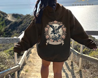 May include: A dark brown hooded sweatshirt featuring a large graphic on the back. The design depicts Saint Michael the Archangel defeating a dragon, surrounded by a floral wreath of roses. The text "St. Michael the Archangel" is printed below.