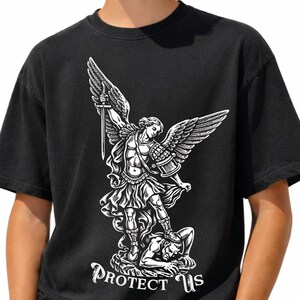 May include: Black t-shirt featuring a white graphic of an angel with a sword, standing over a figure. The text "PROTECT US" is below the graphic. The shirt is made of a soft material.