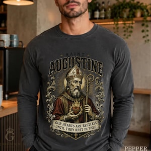 Könnte beinhalten: Ein anthrazitfarbenes Langarmshirt mit einer Grafik von Augustinus von Hippo in Bischofskleidung, der einen Stab und ein brennendes Herz hält. Der Text lautet SAINT AUGUSTINE und OUR HEARTS ARE RESTLESS UNTIL THEY REST IN THEE mit Goldverzierungen.
