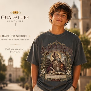 Op de afbeelding: Een antracietgrijs T-shirt met korte mouwen en een grafische print van Sint-Augustinus. Het ontwerp bevat een sierlijk gouden kader, een bisschop, een brandend hart en de tekst "SAINT AUGUSTINE DOCTOR OF THE CHURCH" en "OUR HEARTS ARE RESTLESS UNTIL THEY REST IN YOU".