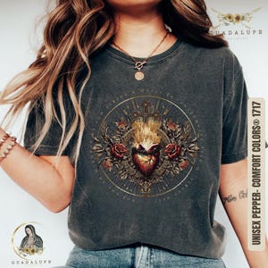 Op de afbeelding: Een donkergrijs T-shirt met een vintage-stijl grafische afbeelding van een hart met vlammen en een kruis omringd door rozen. De afbeelding is in goud en rood met de tekst "IN THE NAME OF THE FATHER AND OF THE SON AND OF THE HOLY SPIRIT" in een cirkel eromheen.