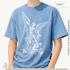 May include: Light blue unisex youth washed denim t-shirt with a white graphic of an angel with a sword, standing over a defeated figure. The text "PROTECT US" is below the graphic. The shirt has short sleeves.