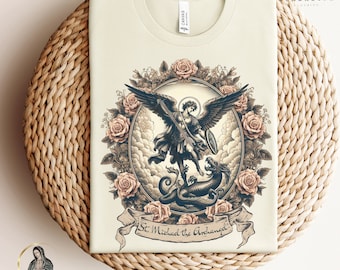 May include: A beige t-shirt with a vintage illustration of Saint Michael the Archangel slaying a dragon. The illustration is surrounded by a wreath of roses and the text "St. Michael the Archangel" is written in a banner below the image.