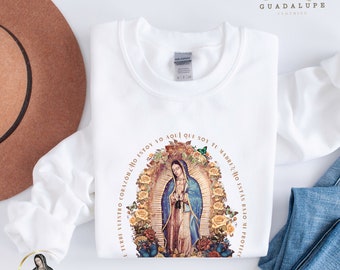 May include: A white long-sleeve sweatshirt featuring a graphic print of Our Lady of Guadalupe surrounded by yellow and red roses and butterflies. The text surrounding the image reads: No se turbe vuestro corazón, ¿no estoy yo aquí que soy tu madre? ¿no estás bajo mi protección?